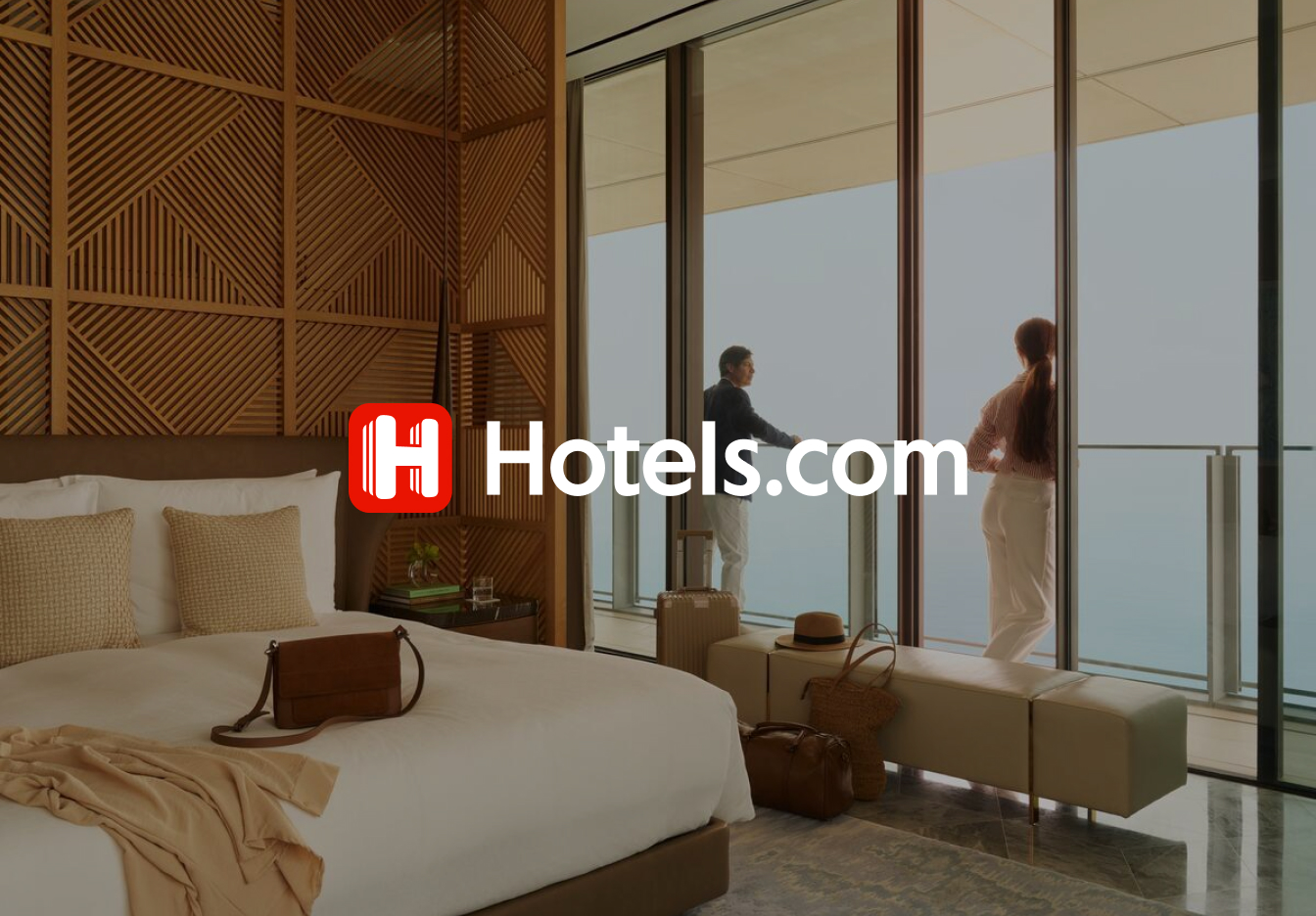 Stylish hotel room with a neatly made bed, warm wood-paneled wall, and travel bags on a bench, with two guests standing on the balcony looking out at the ocean through floor-to-ceiling windows, overlaid with the Hotels.com logo.