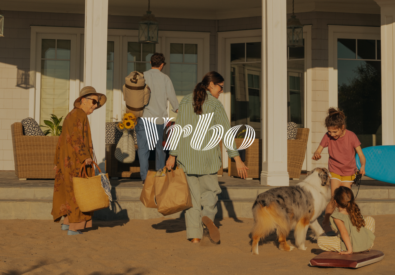 Multi-generational group arriving at a beach house at sunset, carrying bags and sunflowers while children play with a dog in the sandy front yard, with the Vrbo logo overlaid across the scene.