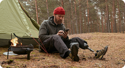 Camper with a prosthetic leg sitting by a tent in the forest, using a smartphone beside a small campfire.