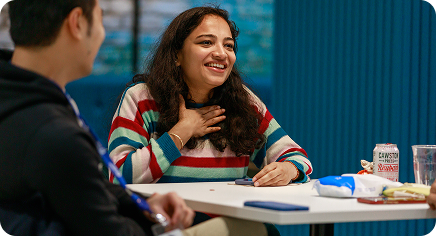 Two colleagues sitting at a table, one smiling and speaking during a casual conversation.