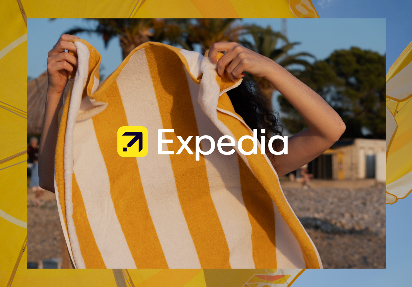 Person on a rocky beach at sunset holding up a large yellow-and-white striped towel that fills the frame, with the Expedia logo centered over the towel and blurred beach huts and palm trees in the background.