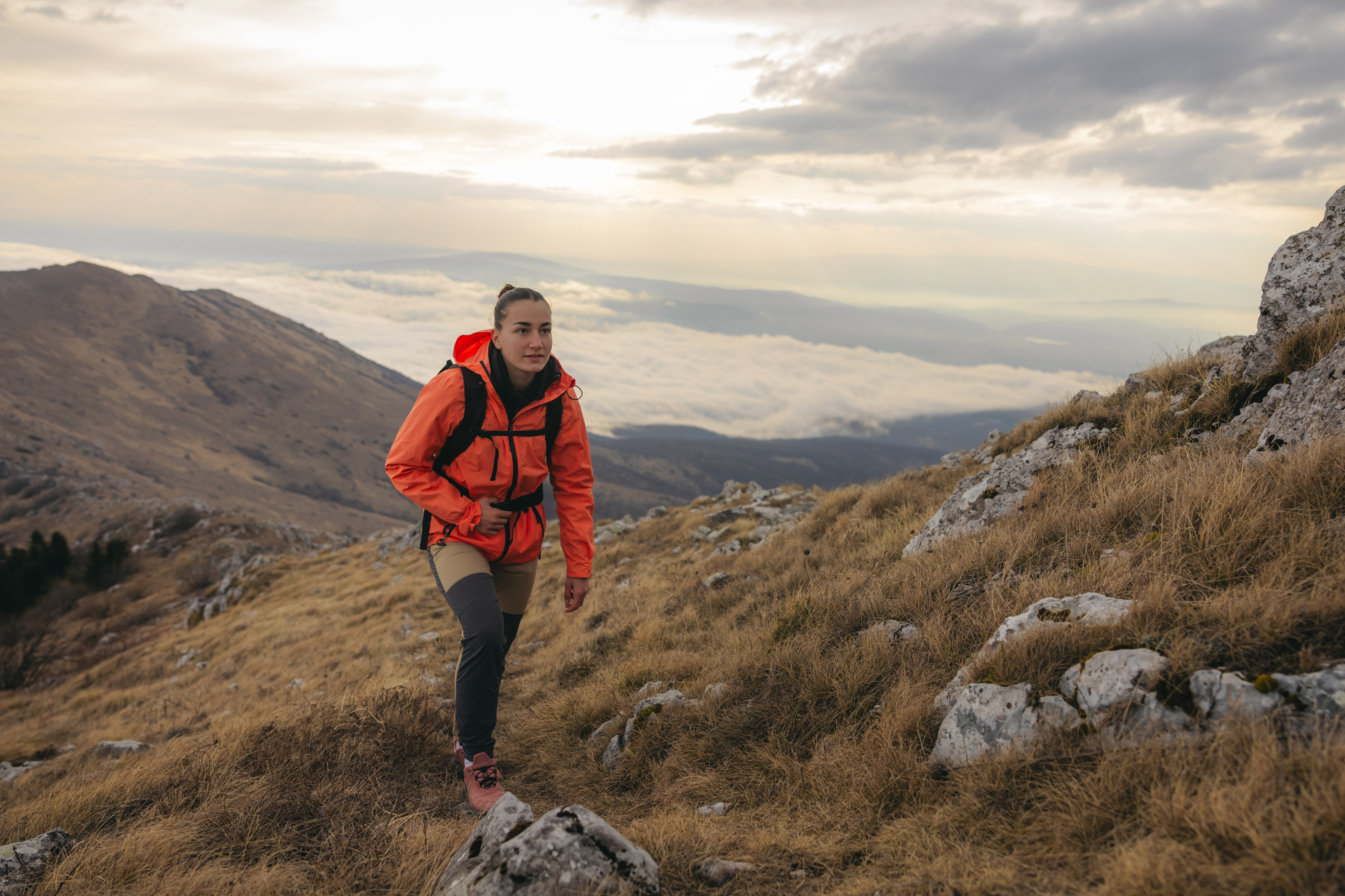 A hiker in an orange jacket climbing a rocky mountainside above the clouds.
