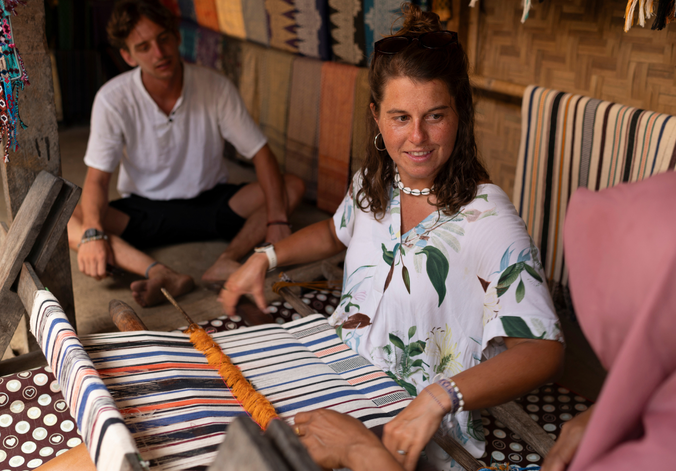 Traveler seated at a traditional handloom weaving a striped textile while smiling and talking with a local instructor just out of frame, with another person sitting on the floor in the background and colorful woven fabrics hanging on the walls of the workshop.